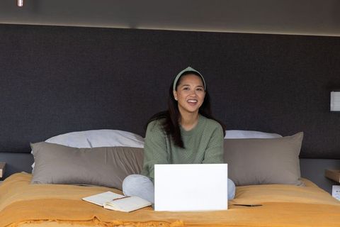 Asian woman sitting cross-legged on bed using laptop and notebook for remote study