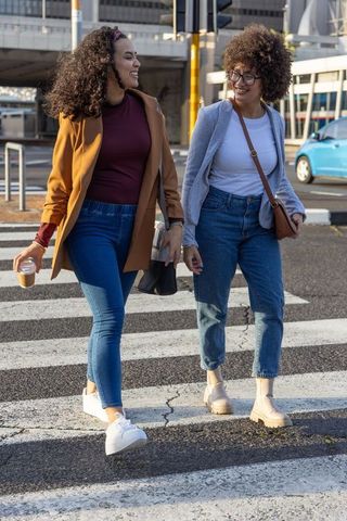 Two african american women walking and laughing on urban crosswalk carrying coffee