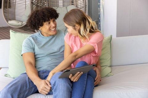 Mixed-race couple cuddling on light sofa sharing tablet on cozy covered patio