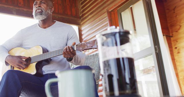 African American man playing acoustic guitar while sitting on porch with French press and coffee cup in foreground. Great for concepts related to leisure, hobbies, music, relaxation, and senior lifestyle. Useful in advertisements promoting coffee brands, senior living communities, or lifestyle blogs about enjoying retirement.