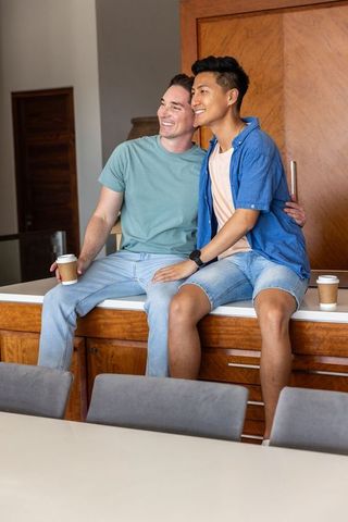 Male friends sitting on kitchen counter holding coffee cups smiling in bright modern home