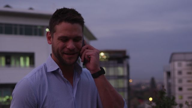 Businessman standing outside office building in evening, talking on smartphone. Useful for concepts like business communication, late work hours, professional lifestyle, or technology in business.