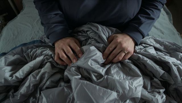 Hands gripping crumpled gray comforter showing wedding ring, man in dark pullover, closeup