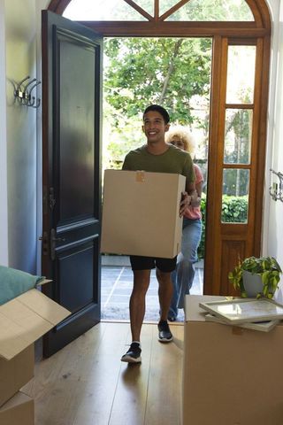Happy diverse couple carrying moving boxes into bright home foyer with wooden door and plants