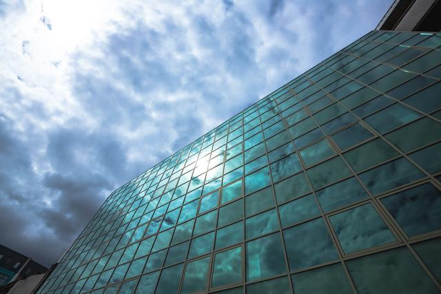 Low angle view of a modern glass skyscraper reflecting the sky and clouds on a sunny day. Ideal for use in business presentations, architectural portfolios, urban development projects, and real estate marketing materials.