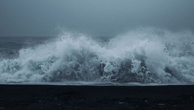 Crashing ocean wave breaking over black sand beach creating dramatic white spray
