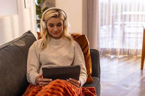 Blonde woman relaxing on sofa with headphones and tablet wrapped in cozy orange blanket