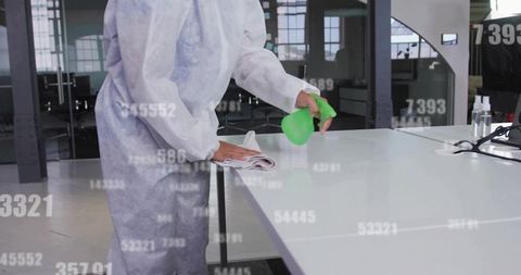 Cleaner in protective coverall disinfecting modern office desk with green spray bottle