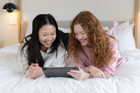 Sharing tablet laughing together on bed relaxing in cozy bedroom for friendship and digital leisure