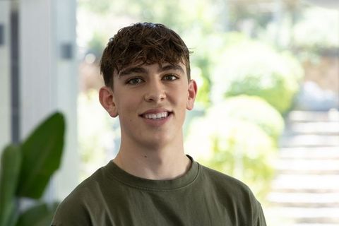 Smiling teenage boy standing by glass door in olive t-shirt with houseplant and sunlight