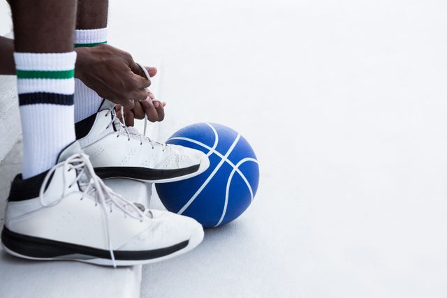 Basketball player tying shoelaces on an outdoor court. Ideal for sportswear advertisements, athletic training materials, fitness blogs, and motivational posters.