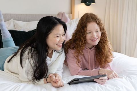 Two friends lying side-by-side on bed watching tablet, cozy home bonding