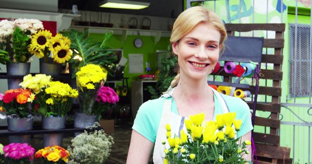Smiling woman holding a bouquet of yellow flowers in a vibrant flower shop. Suitable for representations of florists, retail businesses, flower shops, greeting cards, or advertisements promoting cheerful customer service and professionalism in a local business environment.