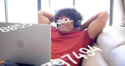 Relaxing man using laptop on sofa, wearing headphones, smiling amid numeric data overlay