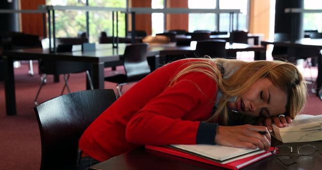 Depicts a student taking a nap while studying in a library. Can be used for themes related to education, study habits, exhaustion, and student life.