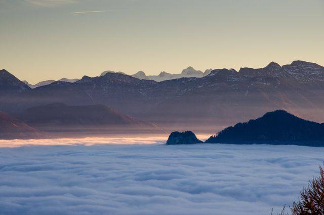 This serene winter scene showcases a sunrise over a cloud-covered valley with towering mountain ranges in the background. Perfect for websites, blogs, and articles about nature, travel, serenity, and winter landscapes. Suitable for promoting outdoor activities and tourism. Works well for prints and screensavers due to its calm and scenic composition.