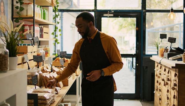 Sunlit grocery shopkeeper stocking shelves wearing mustard shirt and dark apron
