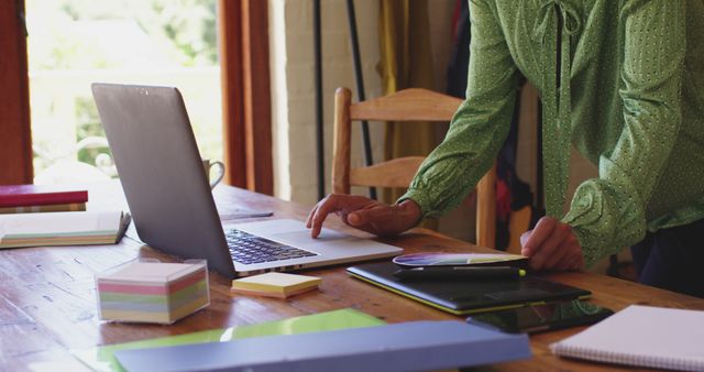 Person standing at desk working from home, using laptop surrounded by documents and office supplies. Ideal for illustrating remote work, home office organization, productivity, and modern work setups in lifestyle blogs, business articles, and productivity guides.