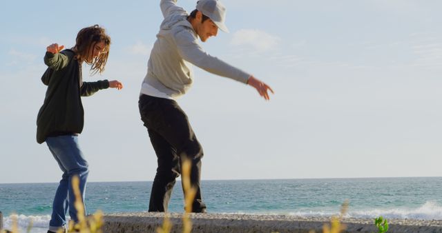 Two friends are balancing on a seawall along the ocean. They are enjoying a sunny day at the beach, wearing casual clothes and showing a carefree attitude. This image can be used in travel promotions, friendship-themed content, and lifestyle blogs to represent joyful and adventurous moments.