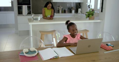 African american mother checking phone while daughter studying on laptop in bright kitchen