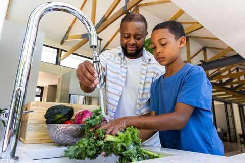 African american father teaching son washing kale in modern kitchen for healthy meals