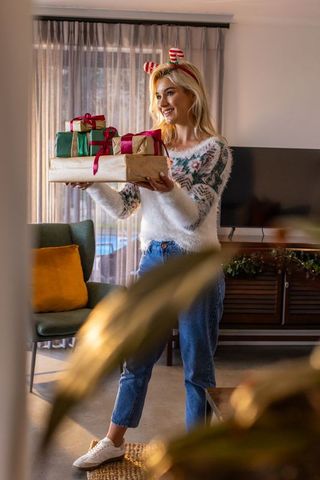 Young woman wearing festive headband holding wrapped christmas gifts in cozy living room