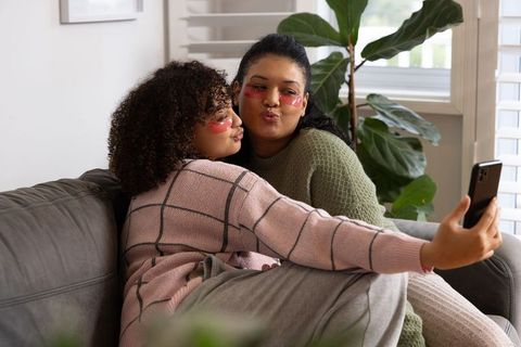 Multicultural friends posing for selfie on couch wearing pink under-eye patches, cozy self-care mome