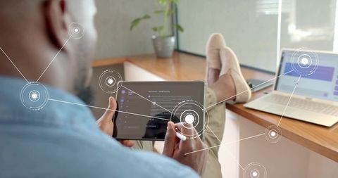 Man Using Tablet with Stylus at Sunlit Home Workspace with Laptop and Digital HUD Overlays