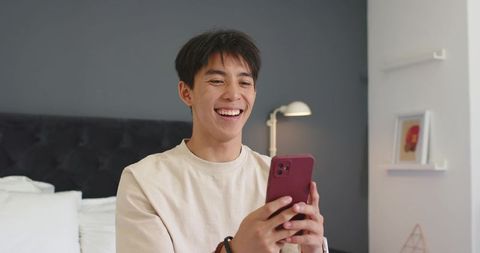 Smiling asian man holding smartphone in modern bedroom receiving app notification