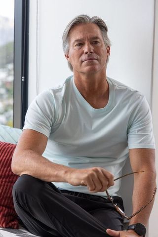 Mature man sitting by window holding eyeglasses in minimalist apartment, serene portrait