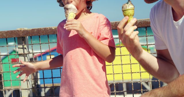 Child and adult enjoying ice cream cones on a sunny day at the beach. Bright, colorful beach huts visible in the background. Great for themes of summer, family bonding, outdoor activities, and refreshing treats. Perfect for advertising summer vacations, family-oriented products, or beach-related activities.