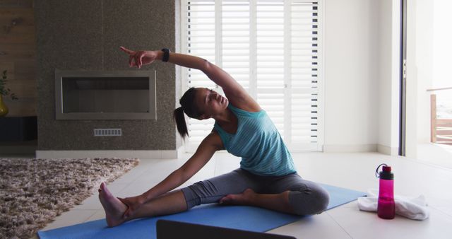 Woman practicing yoga in living room following online class on laptop. She is stretching on a yoga mat, with a water bottle nearby. Ideal for illustrating home fitness routines, online workout classes, and promoting a healthy lifestyle.