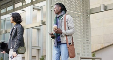 African American commuter standing holding coffee and smartphone at modern transit entrance