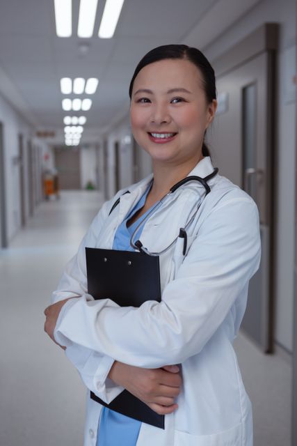 Female doctor standing in a hospital corridor, holding a clipboard and smiling confidently. Ideal for use in healthcare advertisements, medical articles, hospital brochures, and websites promoting medical services.