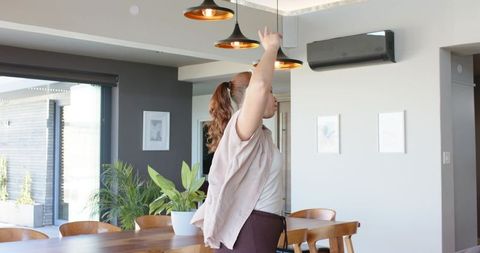 Woman reaching up in modern sunlit dining room with pendant lights and indoor plants