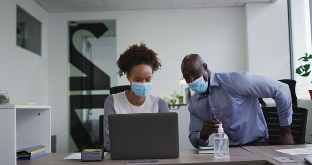 Two business colleagues in office wearing face masks, reflecting safety measures during the COVID-19 pandemic. Man and woman working together on project using laptop, hand sanitizer in view. Ideal for topics on workplace safety, office collaboration, pandemic protocols, or corporate lifestyle.