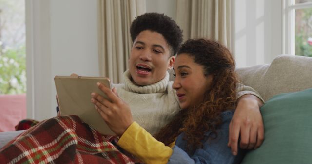 Image of happy biracial couple siting on sofa under blanket and using tablet. Domestic lifestyle and leisure time at home.