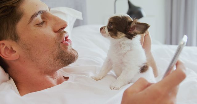 Man affectionately interacting with small puppy while holding smartphone on bed. This can be used for themes related to pet care, human-animal bond, relaxation, modern lifestyle, and communication. Great for blogs, articles, or advertisements about pets, technology, or home life.