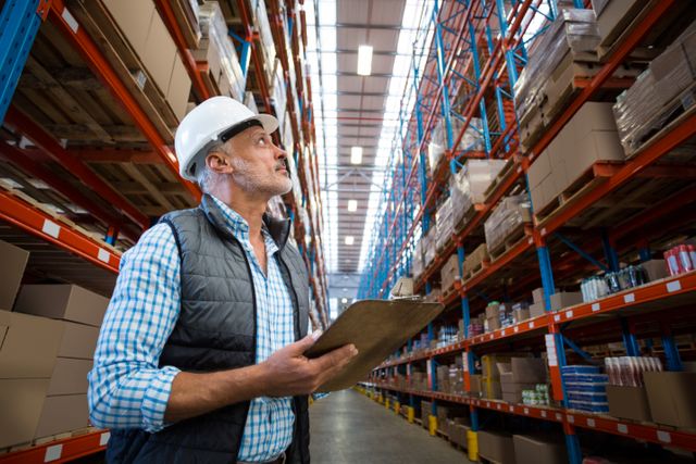 Warehouse worker inspecting inventory with clipboard, wearing a hard hat and safety vest. Ideal for use in articles or advertisements related to logistics, supply chain management, industrial safety, and warehouse operations. Can also be used for illustrating topics on inventory management, storage solutions, and distribution centers.