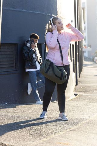 Young woman adjusting headphones while young man talking on phone by sunlit urban wall