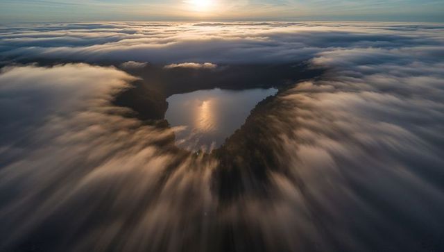 Sunrise fog pouring around circular forest lake, sun reflecting on calm mirror water