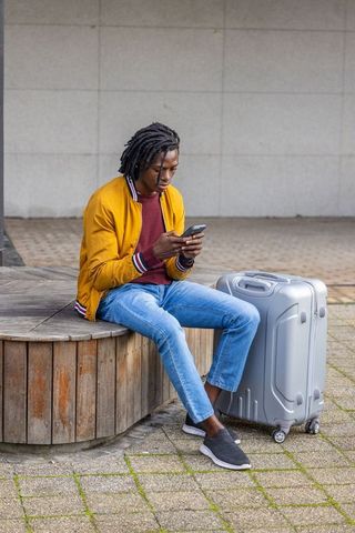 African american traveler sitting on bench checking phone with suitcase in urban plaza