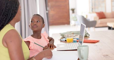 African mother guiding daughter homeschooling at kitchen table with laptop and juice