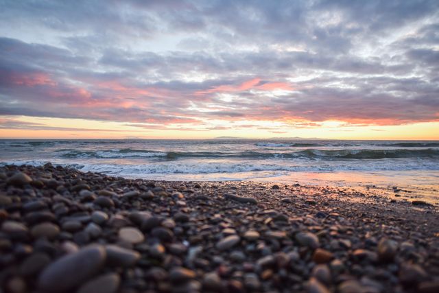 Sun setting over the ocean with clouds filling the sky. Gentle waves touch the rocky pebble beach with vibrant sunset colors reflecting on the water. Perfect for travel blogs, relaxation visuals, marine life articles, and sunset-themed designs.