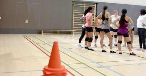 Diverse female volleyball team training with coach on indoor hardwood court near cones