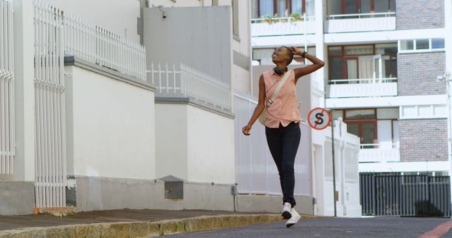 Young African woman walking confidently in an urban area wearing headphones, expressing a carefree and modern lifestyle. This image can be used for urban living concepts, fashion, self-confidence themes, or technology integration in everyday life.