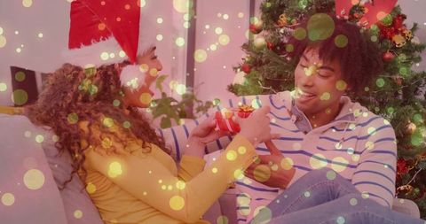Couple exchanging christmas gift on couch by decorated tree with cozy bokeh lights