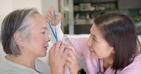 Home caregiver adjusting oxygen mask on senior woman, nursing aide assisting breathing