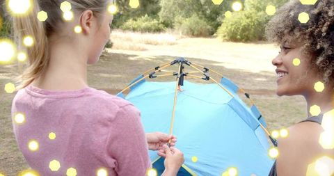 Two women assembling blue tent in sunny meadow, smiling and collaborating on camping setup