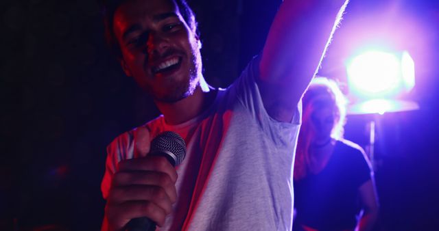 A young Caucasian man is singing into a microphone with a joyful expression, illuminated by colorful stage lights, with copy space. His performance exudes confidence and entertainment value, capturing a moment of musical expression.